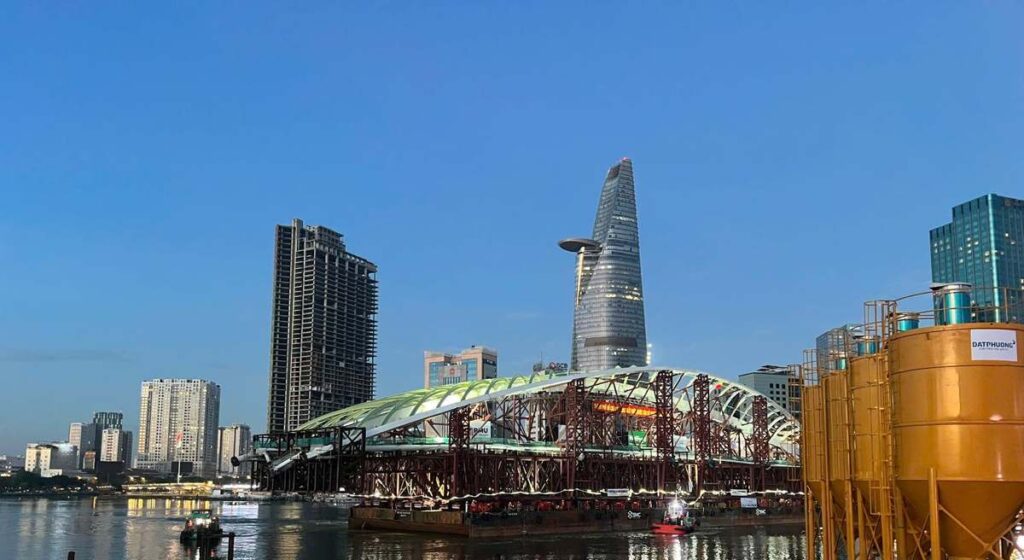 Nhịp vòm cầu đi bộ qua sông Sài Gòn lúc chạng vạng trước giờ hợp long, phía sau là trung tâm Quận 1 cũ | Saigon River pedestrian bridge arch at dusk before closure, District 1 skyline behind